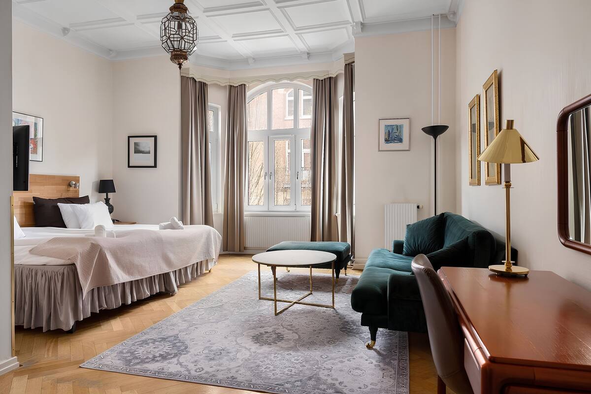 A hotel room with a bed, a green sofa, a wooden desk, a round table, and a large window with curtains.