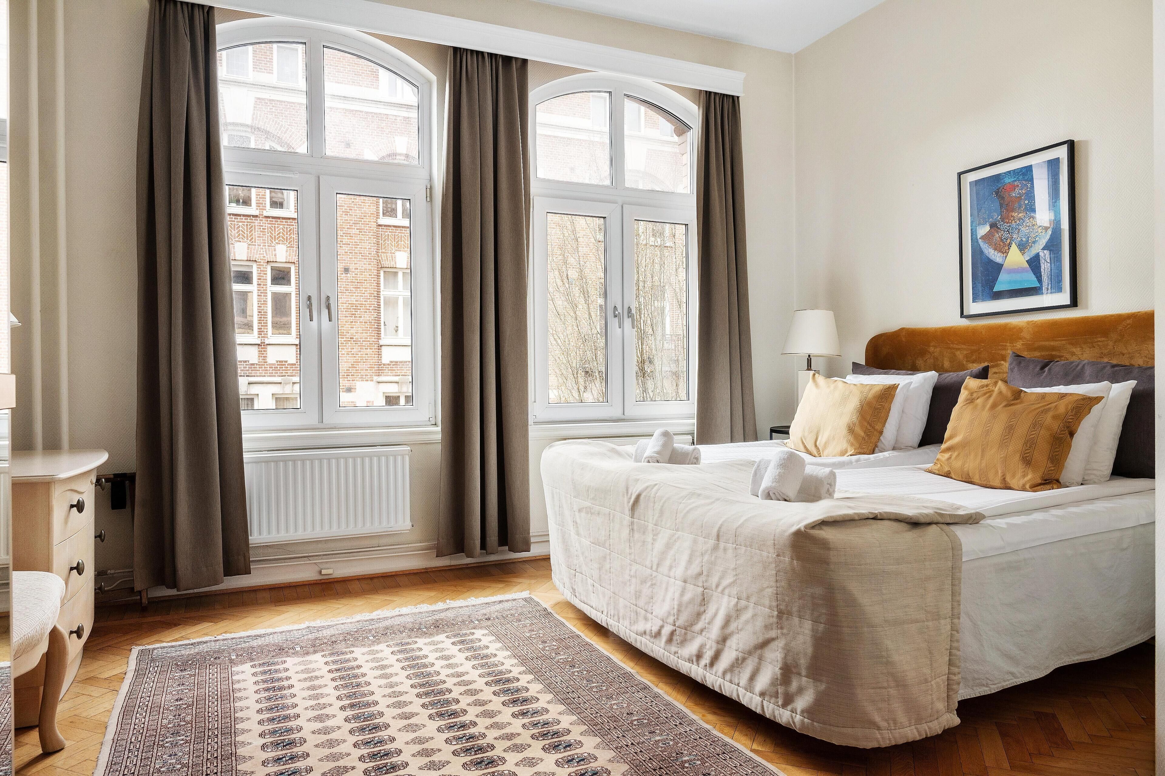 Une chambre avec une grande fenêtre, un lit recouvert de plusieurs coussins, une table de chevet avec une lampe et un tapis sur le sol.