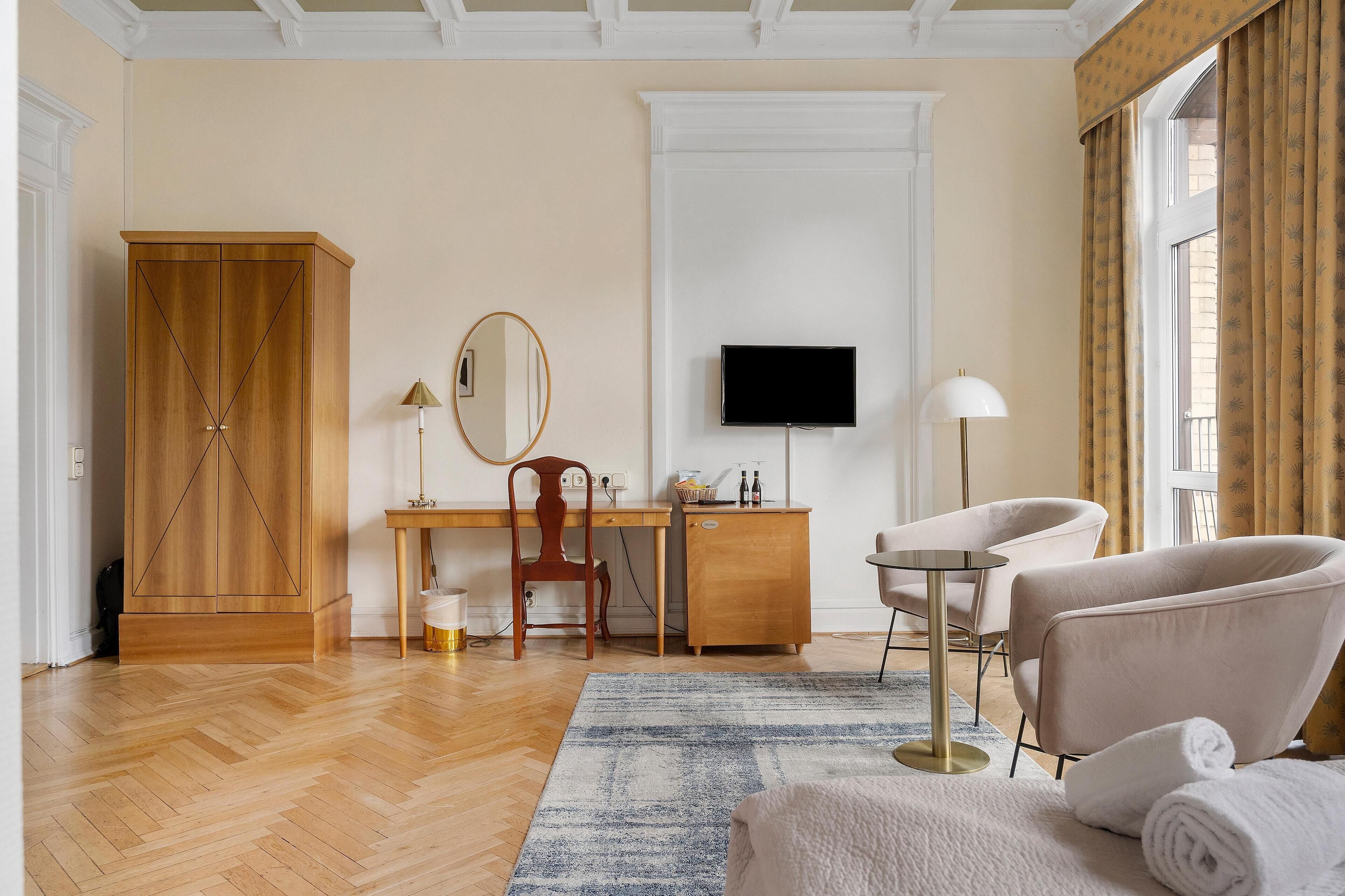 A room with a wooden wardrobe, a desk with a chair, a television, a lamp, and a sofa with a rolled-up towel.