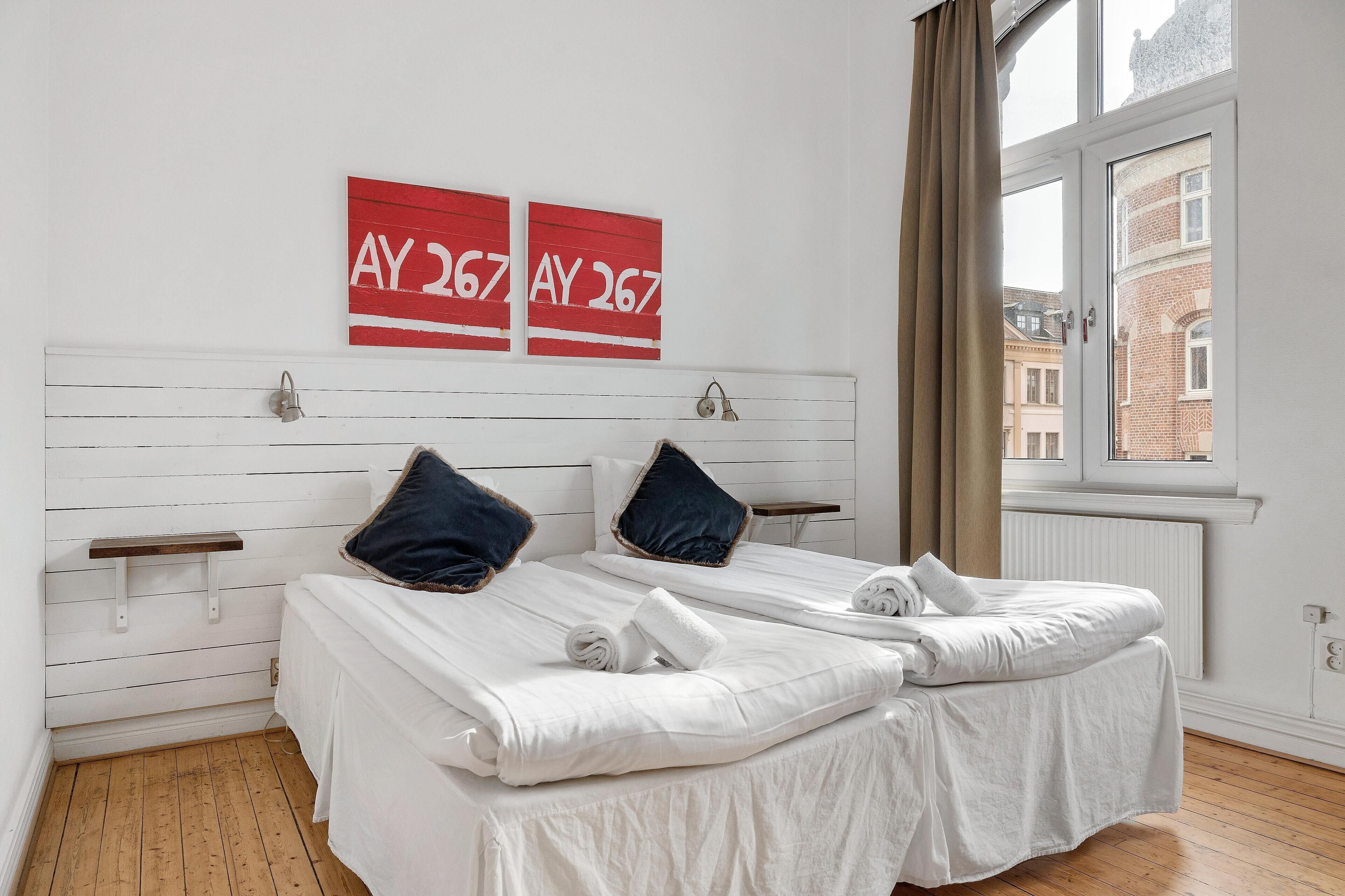 Een tweepersoonskamer met twee bedden, een raam met uitzicht op gebouwen en een rood bord met 'AY 267' aan de muur.