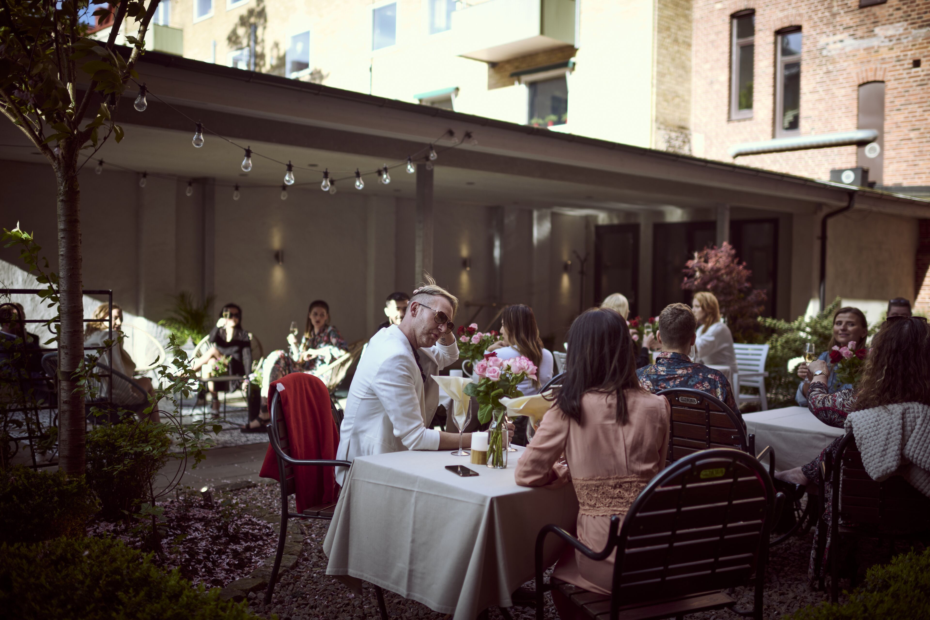 Un área de comedor al aire libre con mesas y sillas, rodeada de follaje y edificios al fondo.