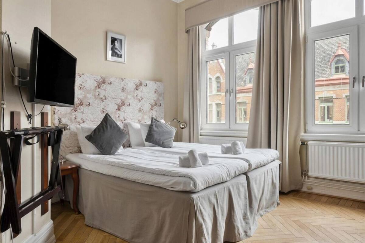 A neatly made bed with pillows and towels, a television mounted on the wall, and a window with curtains.