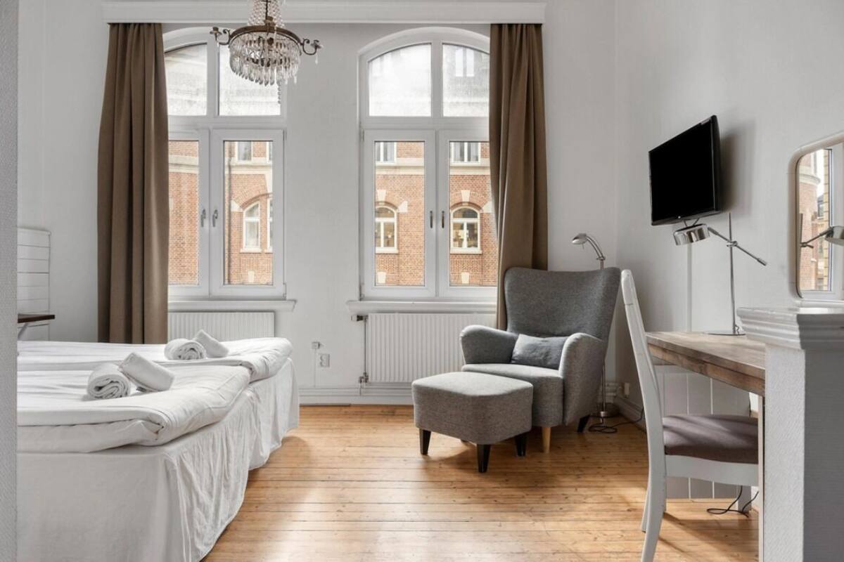 A hotel room with a bed, a desk, a chair, a TV, and large windows with curtains.