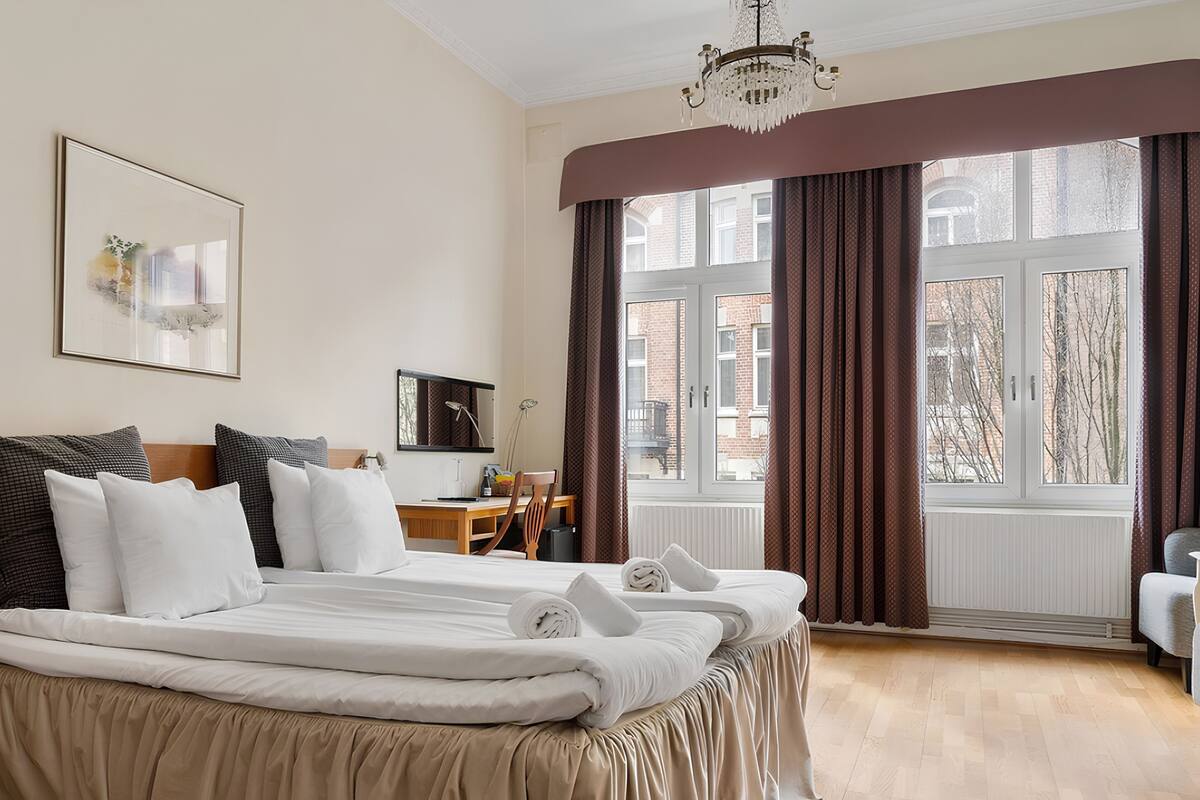 A hotel room with a large bed, a desk, a chair, and a view of the city through large windows.