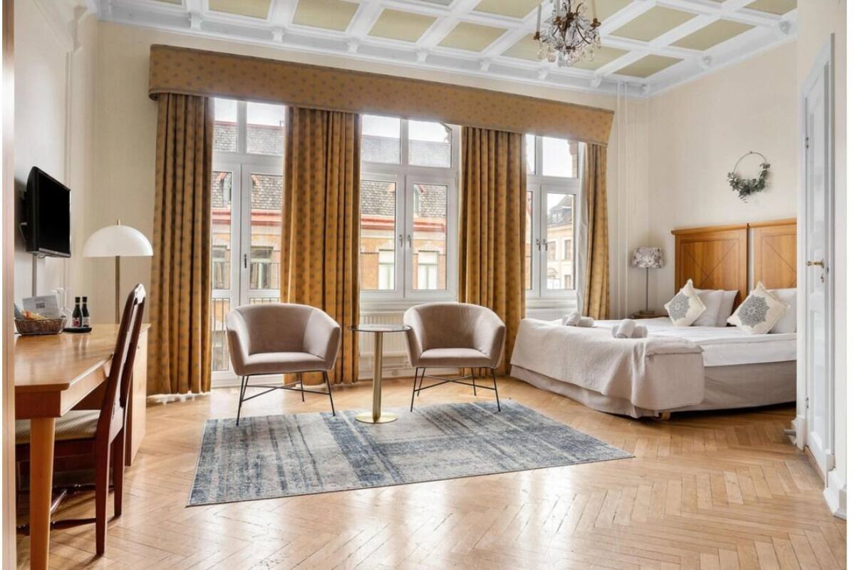 A hotel room with a bed, two armchairs, a small table, a TV, and large windows with curtains.