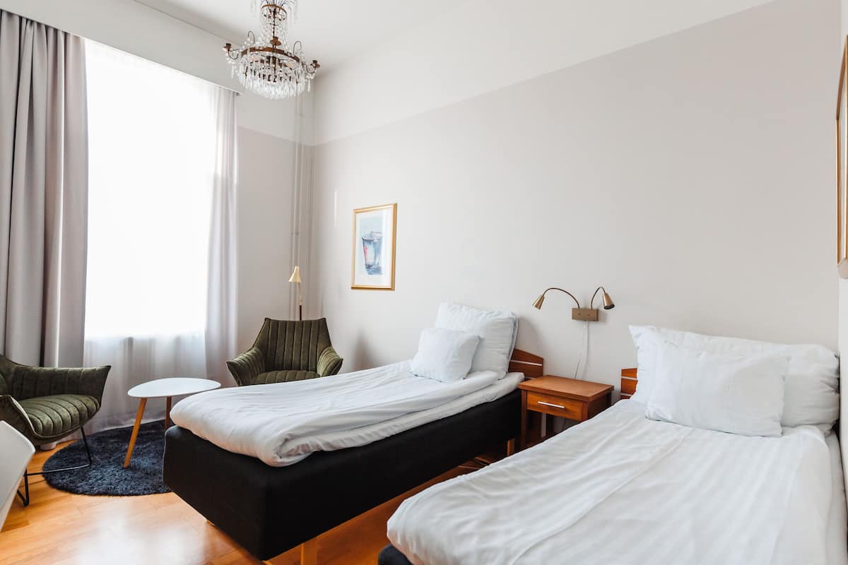 Hotellihuone, jossa on kaksi sänkyä, pieni pöytä, tuoli, kattokruunu ja ikkuna, jossa on verhot.