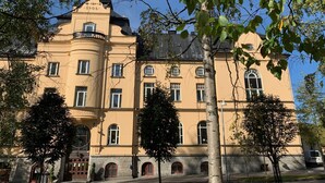 Un grand bâtiment historique à la façade jaune, doté de nombreuses fenêtres et d’une tour imposante sur laquelle est inscrite l’année 1906.