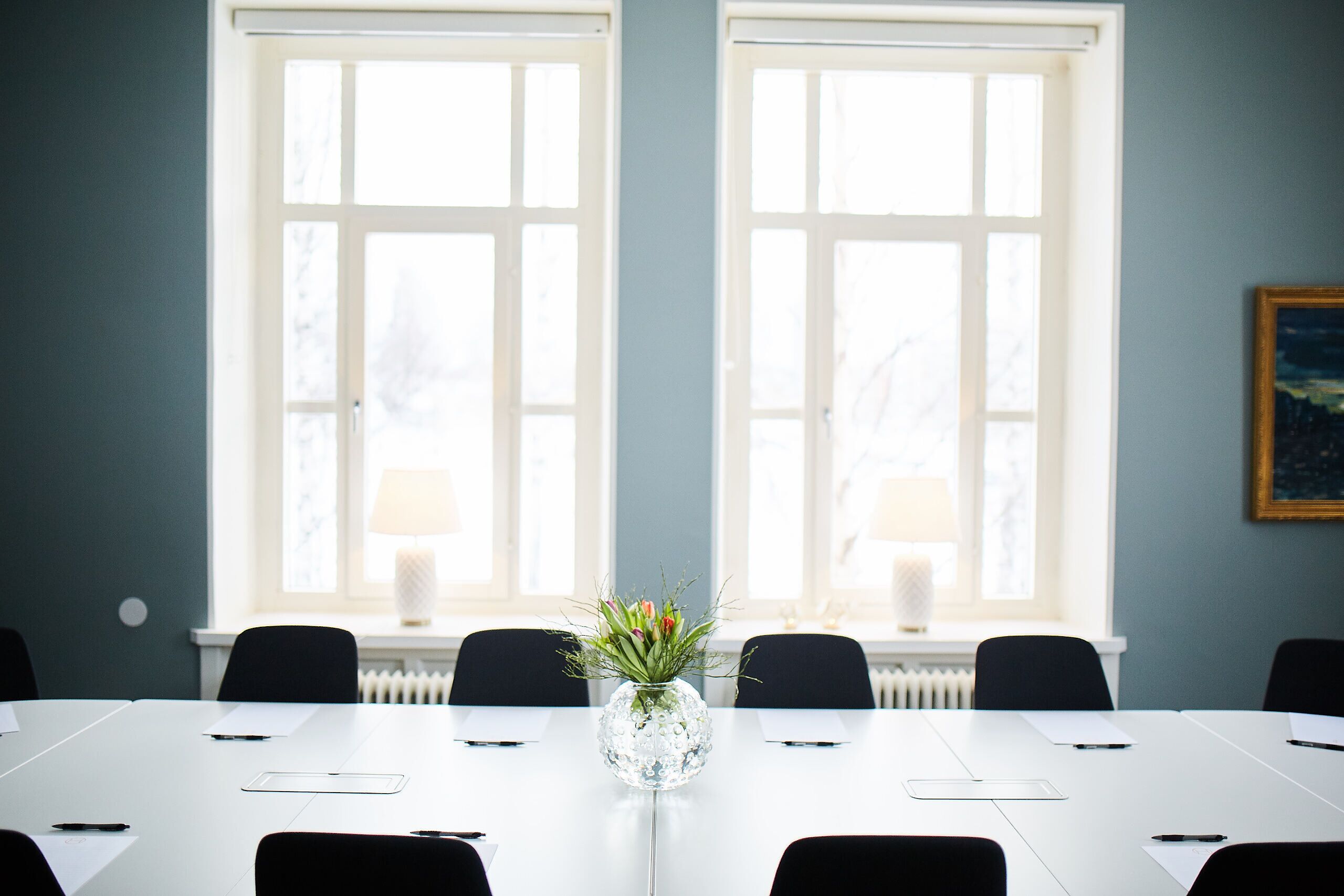 Un salón de conferencias con una mesa blanca, sillas negras, un jarrón de cristal con flores y dos ventanas con persianas.