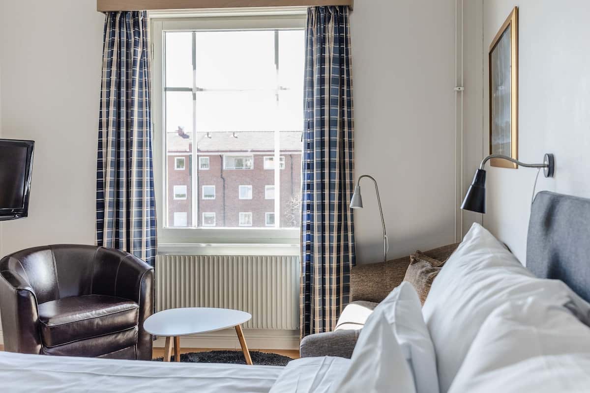 Hotellihuone, jossa on sänky, nahkainen nojatuoli, pieni pöytä, sohva ja ikkuna, jossa on verhot.
