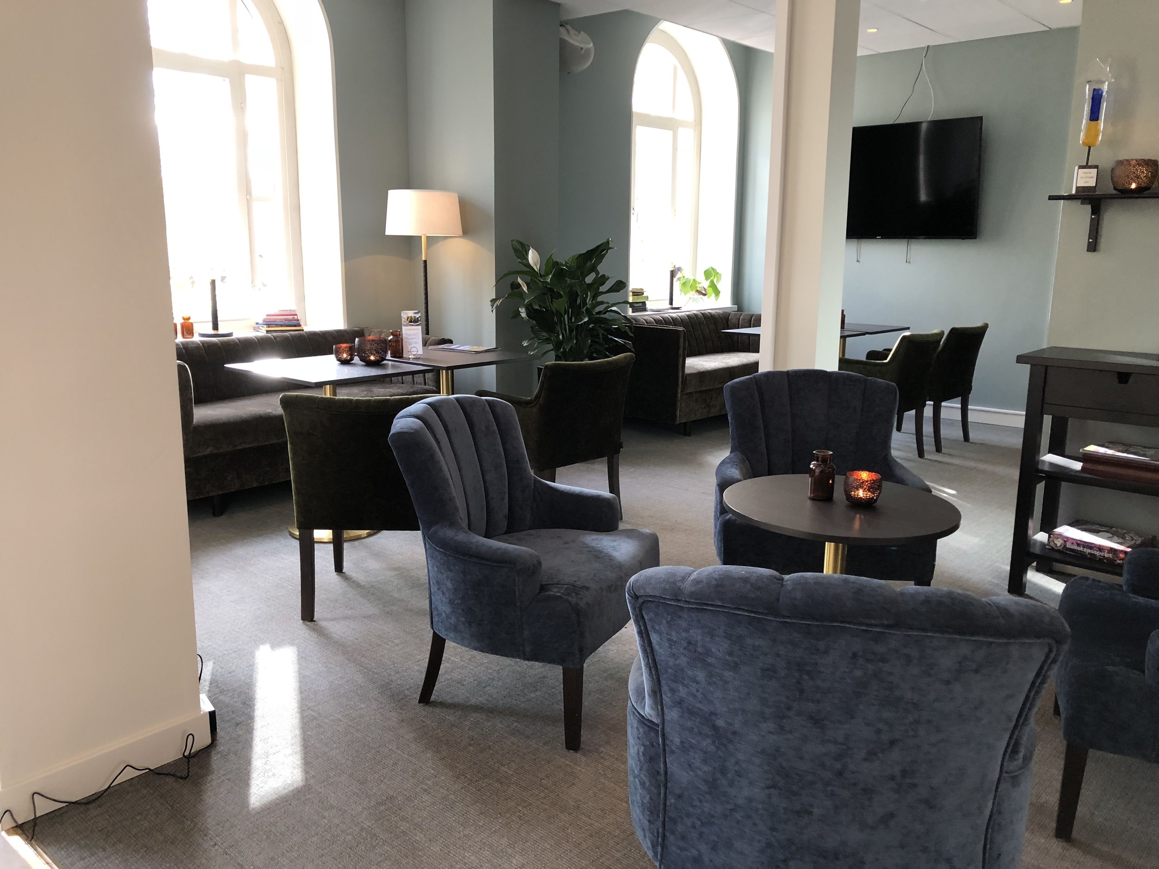 A modern hotel lobby with a sofa, armchairs, a dining area, and a TV.
