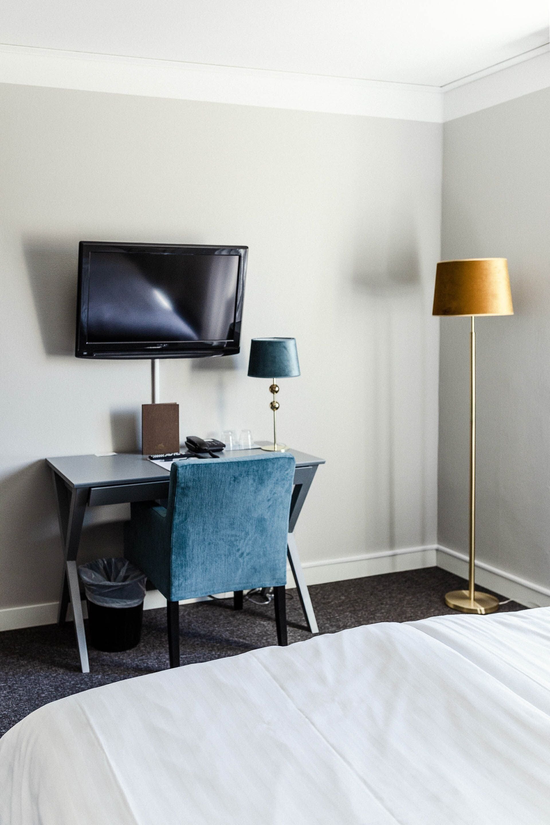 A hotel room with a bed, a desk with a computer, a television, a lamp, and a trash bin.