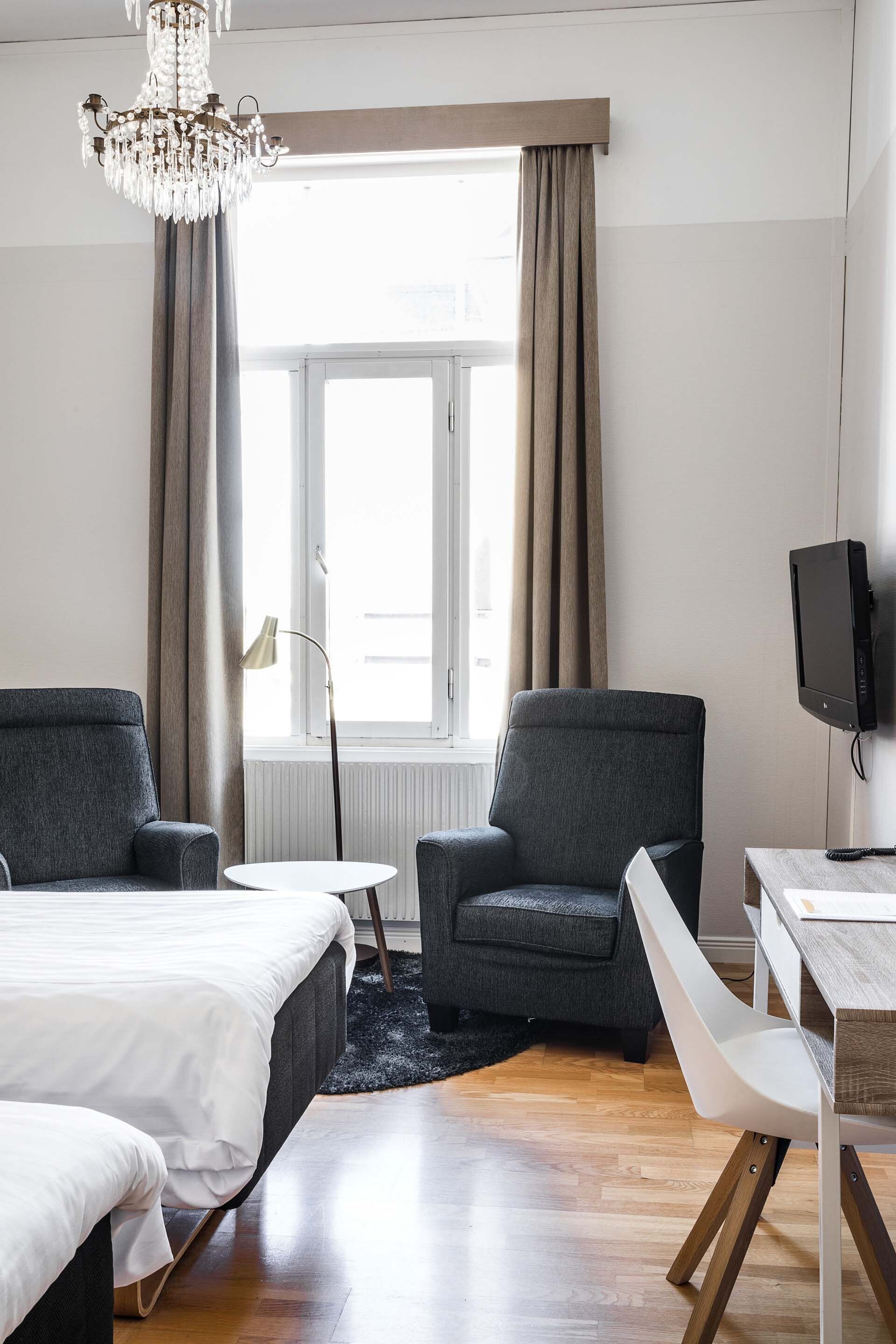 A hotel room with a bed, two armchairs, a small table, a TV, and a large window with curtains.