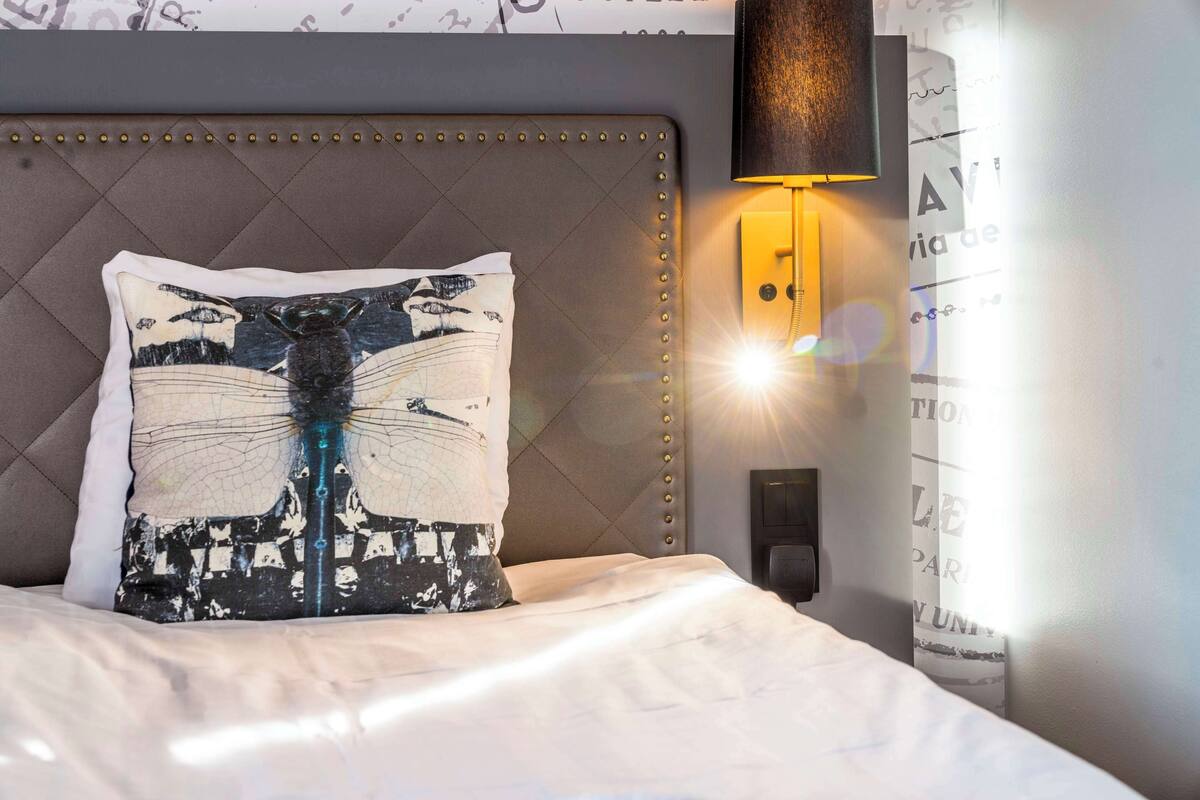 A neatly made bed with a decorative pillow, a wall-mounted light, and a partial view of a poster or sign.