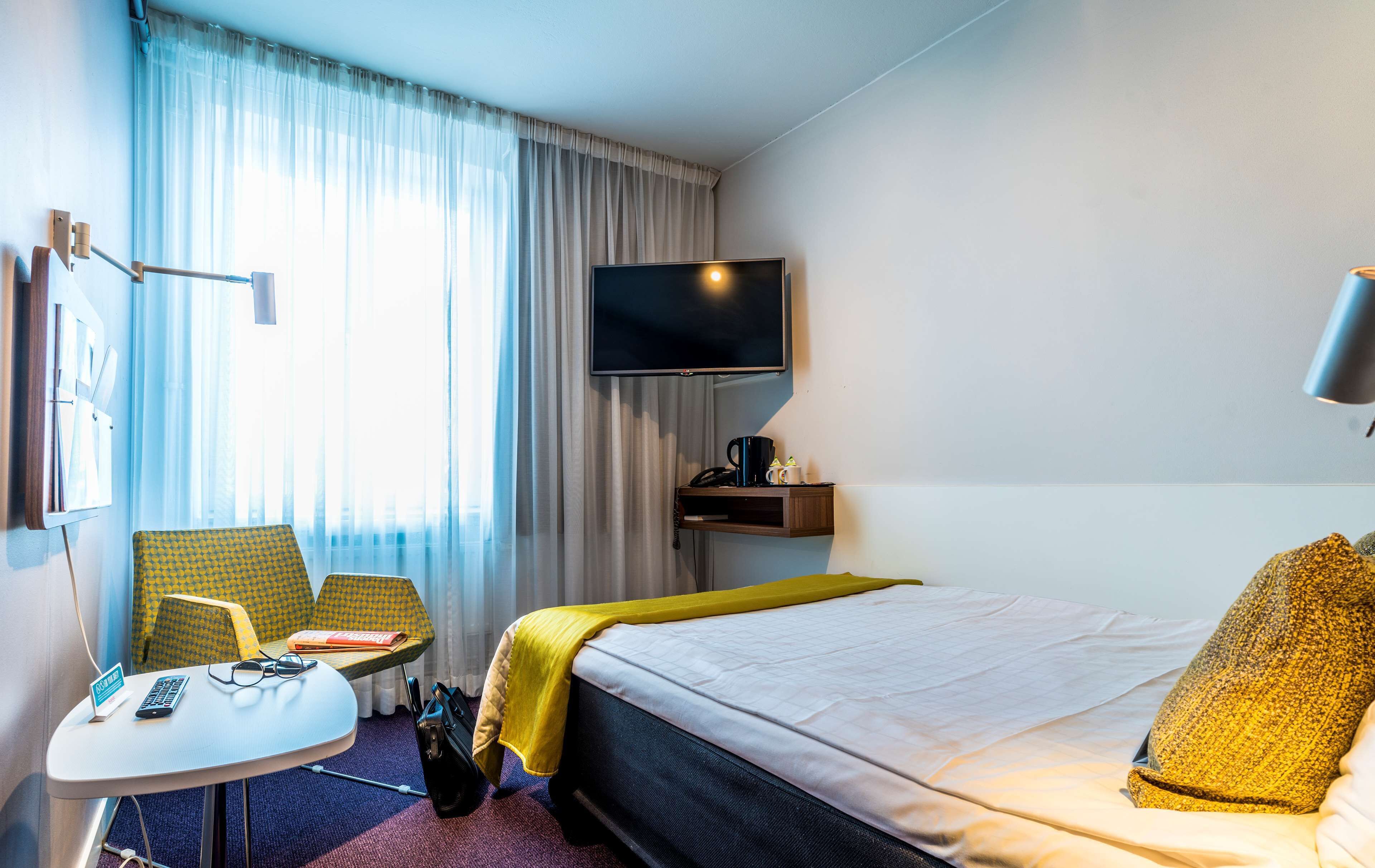A hotel room with a bed, a desk with a chair, a TV, and a window with curtains.