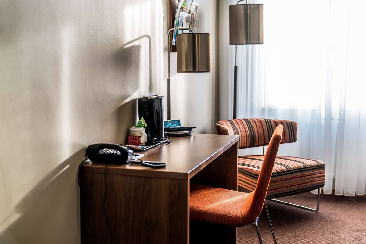 A hotel room with a wooden desk, a striped armchair, a standing lamp, and a window with sheer curtains.