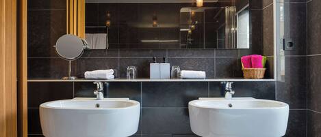 Une salle de bain moderne avec deux lavabos blancs, un miroir et une étagère où sont posées des serviettes et un panier.