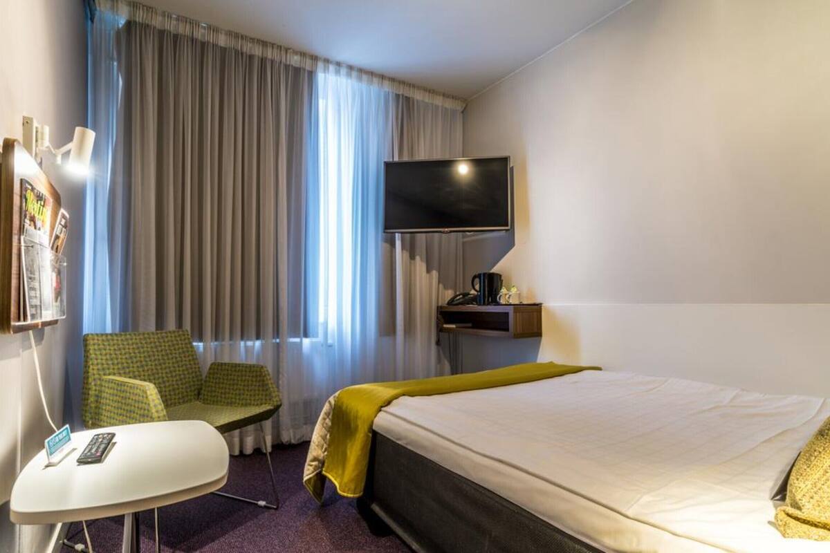 A hotel room with a bed, a TV mounted on the wall, a small table with a remote control, a chair, and a window with curtains.