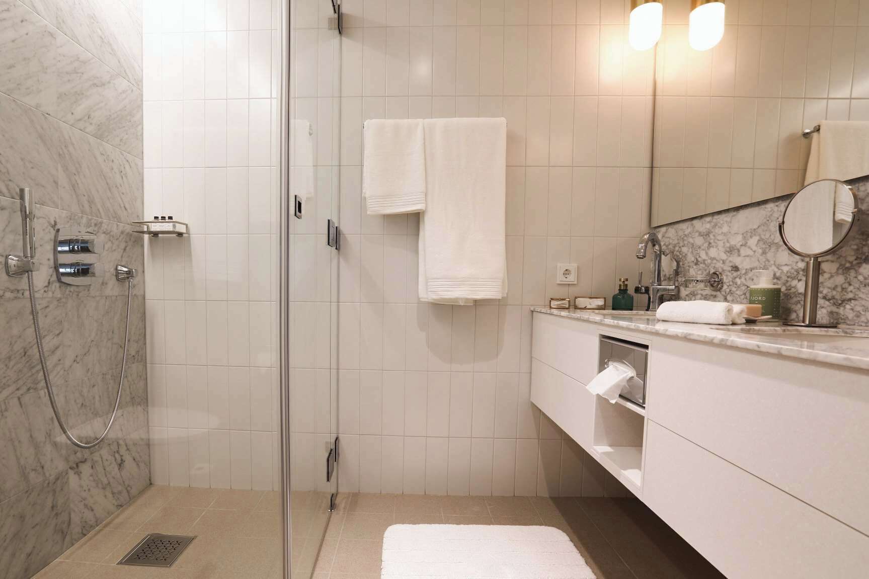 Un baño moderno con ducha envuelta en vidrio, paredes de azulejos blancos, encimera de mármol y un espejo grande.