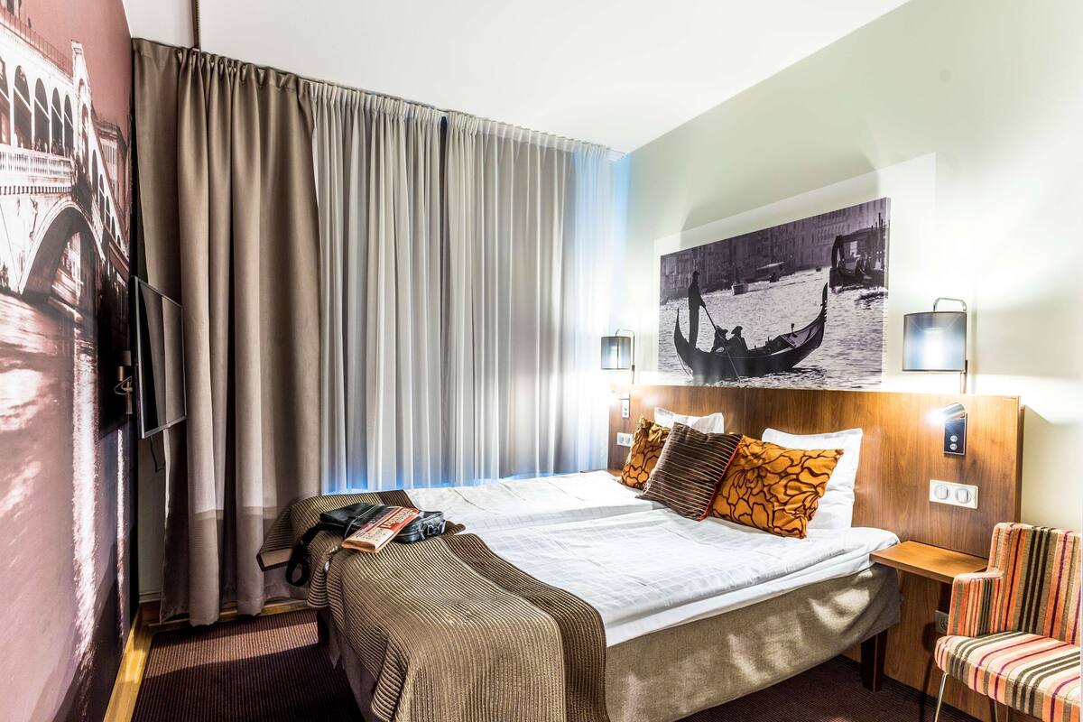 A hotel room with a bed, a chair, a desk, a TV, and a wall with a picture.