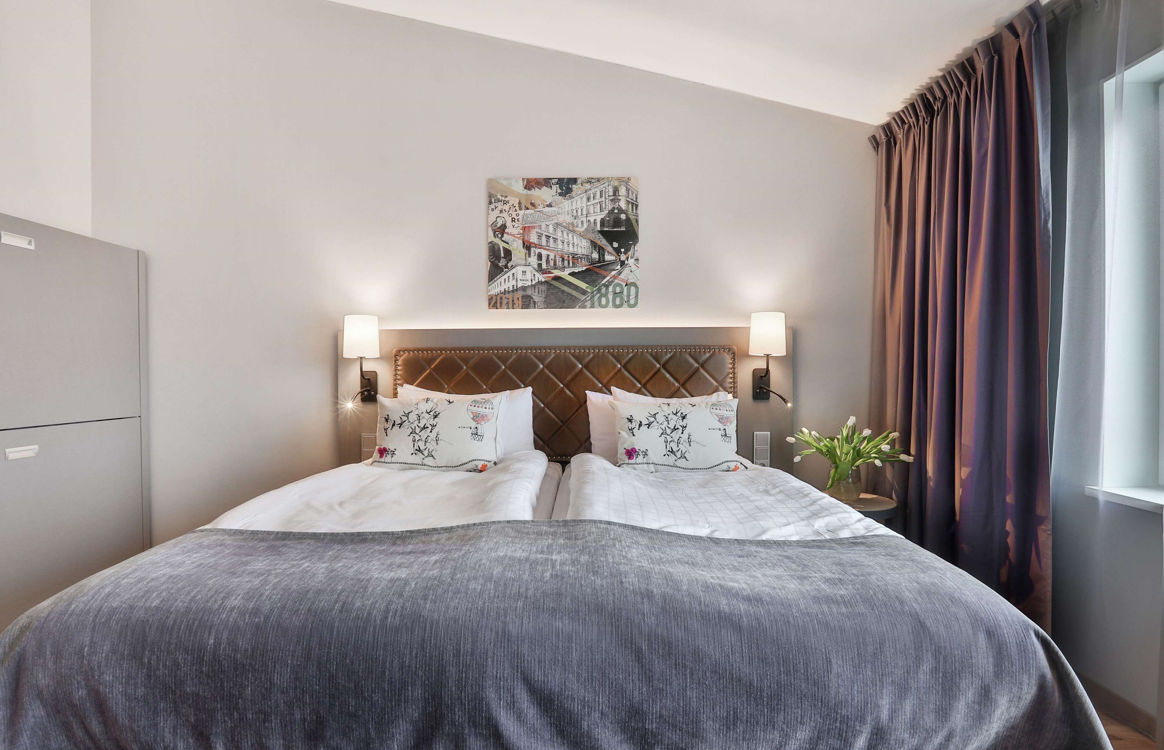 Camera d'albergo con un letto, due abat-jour, una testiera con un'opera d'arte che raffigura un panorama urbano, una finestra con tende e una piccola pianta sul comodino.