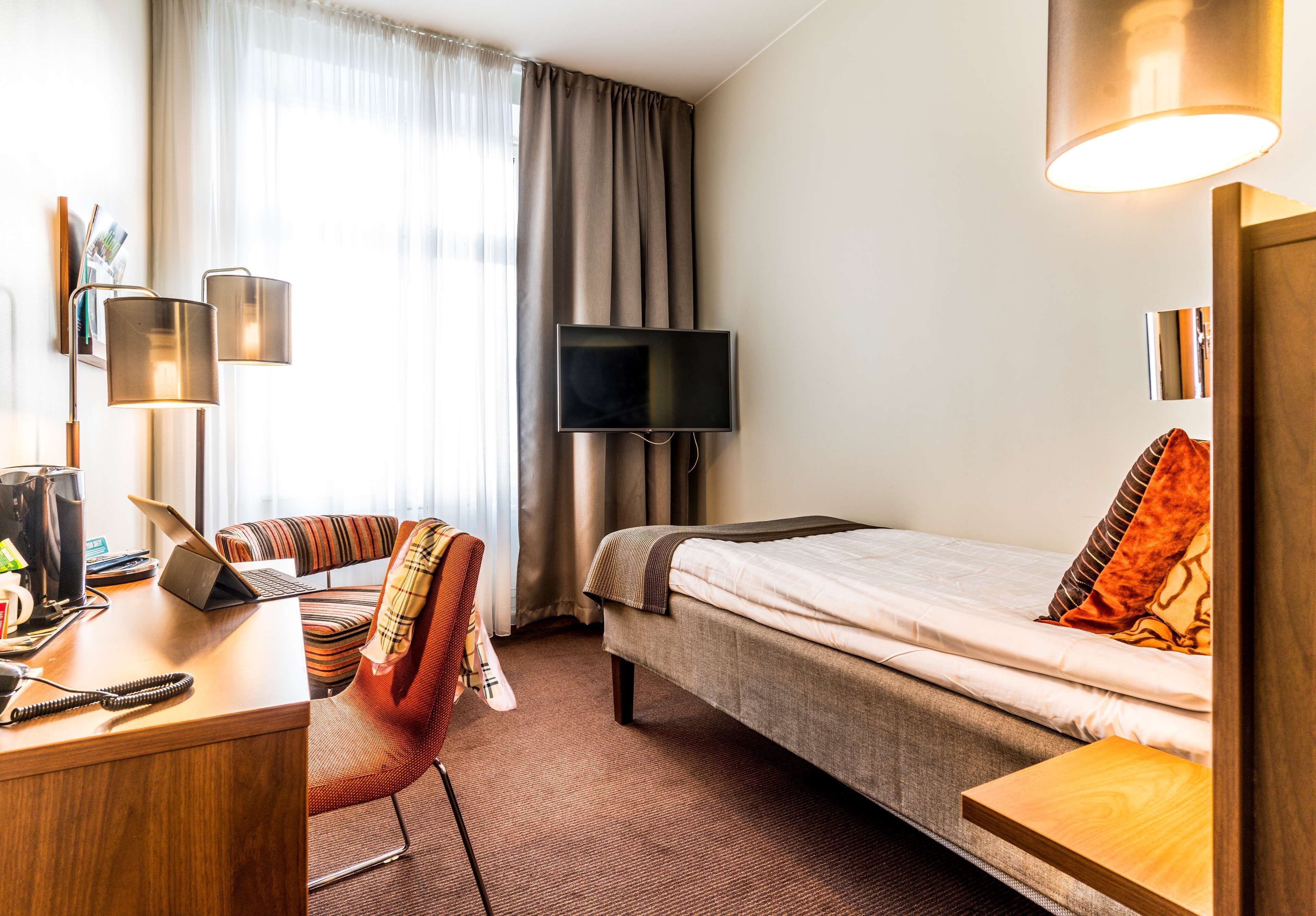 A hotel room with a bed, a desk with a laptop, a chair, a television, and a lamp.
