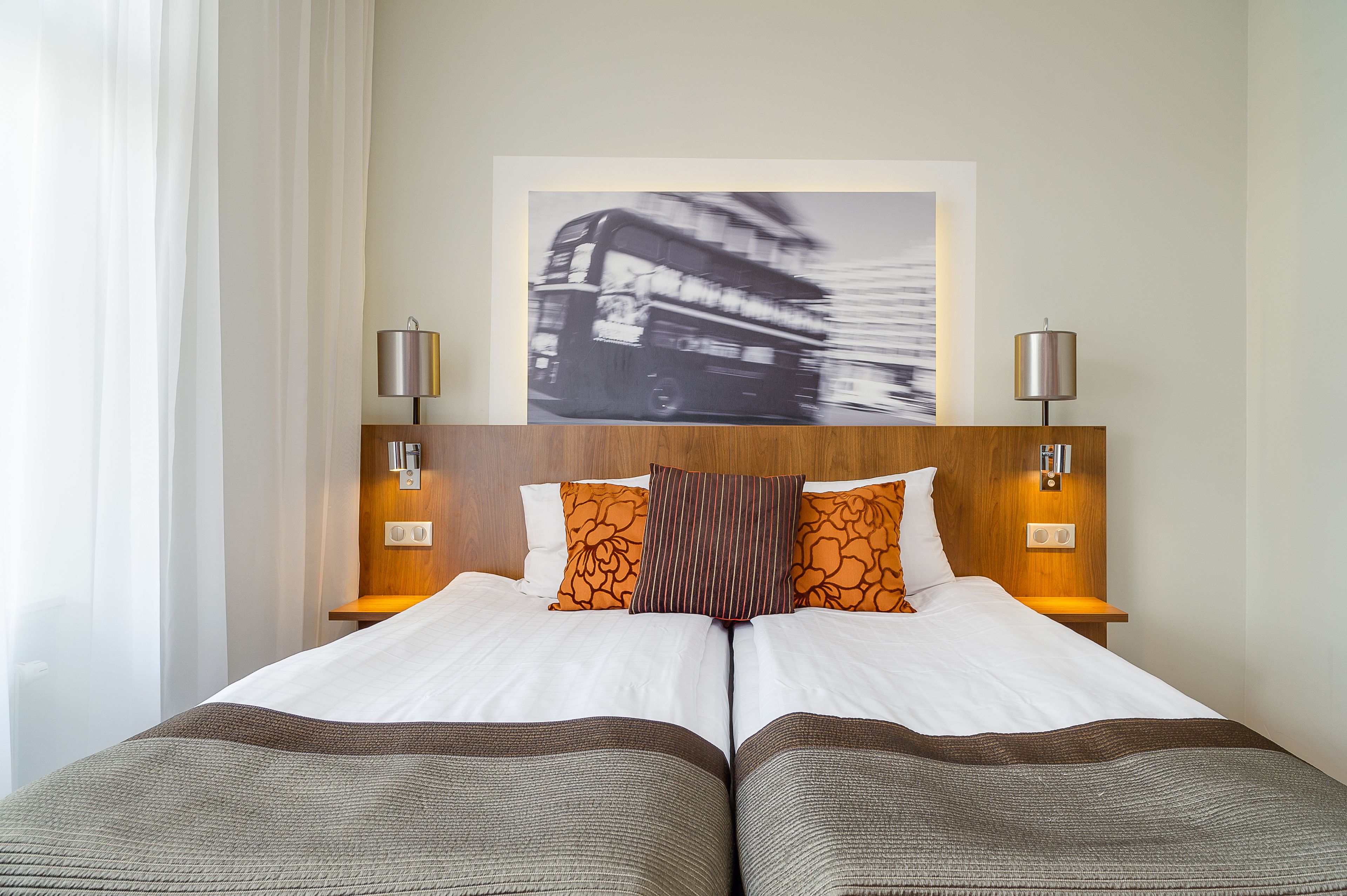 Una cama doble con ropa de cama blanca y marrón, dos lámparas de noche, un cabecero de madera y una obra de arte de un autobús en blanco y negro sobre la cama.