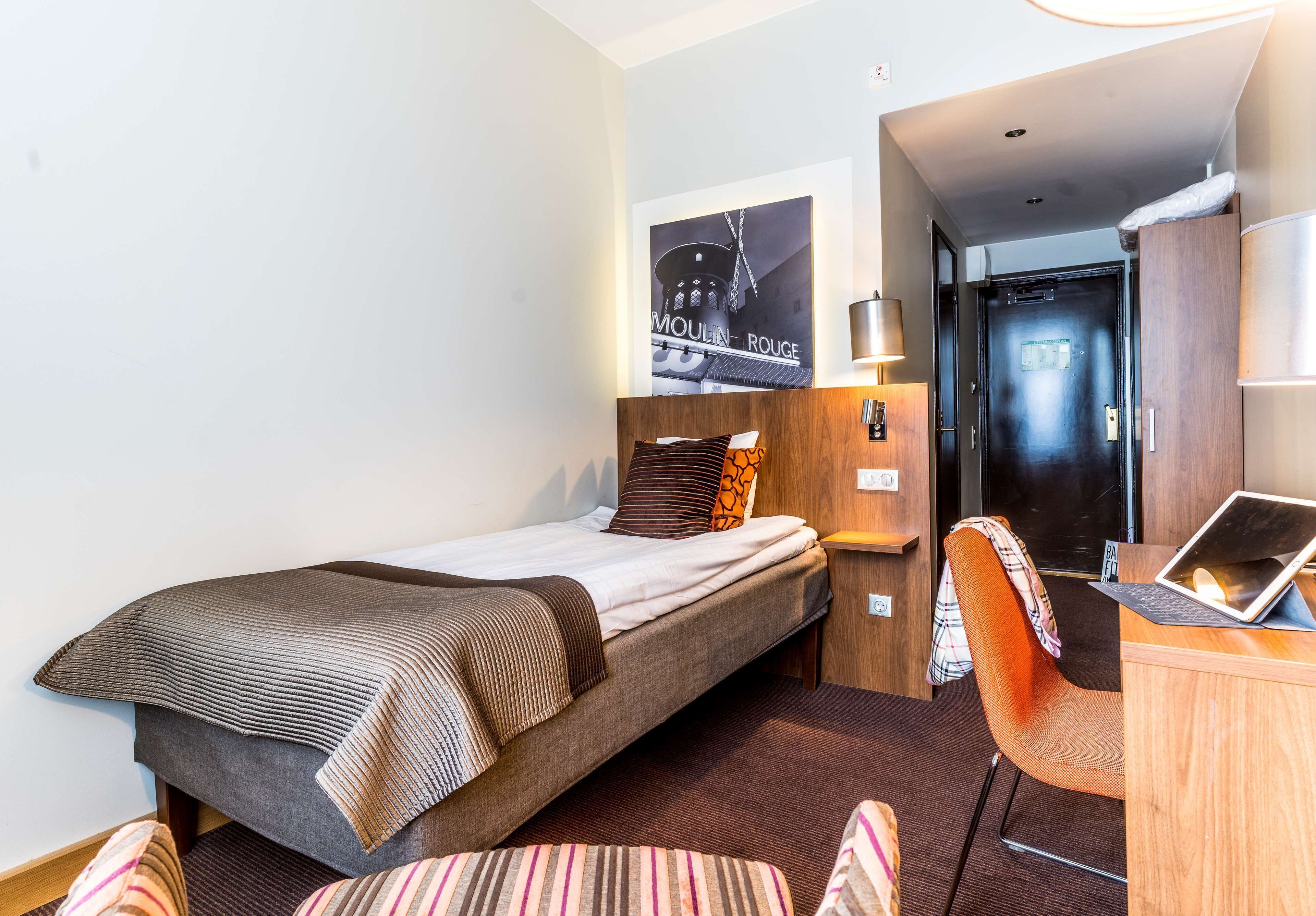 Quarto de hotel com uma cama, uma escrivaninha com um laptop, uma cadeira e uma luminária na parede.