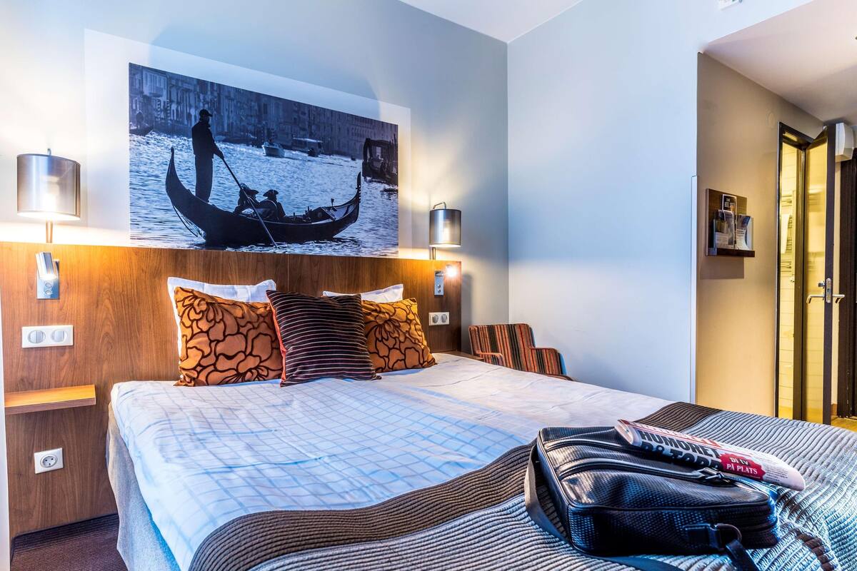 A hotel room with a bed, a wooden headboard, a painting of a gondola, two bedside lamps, a nightstand, and a suitcase on the bed.