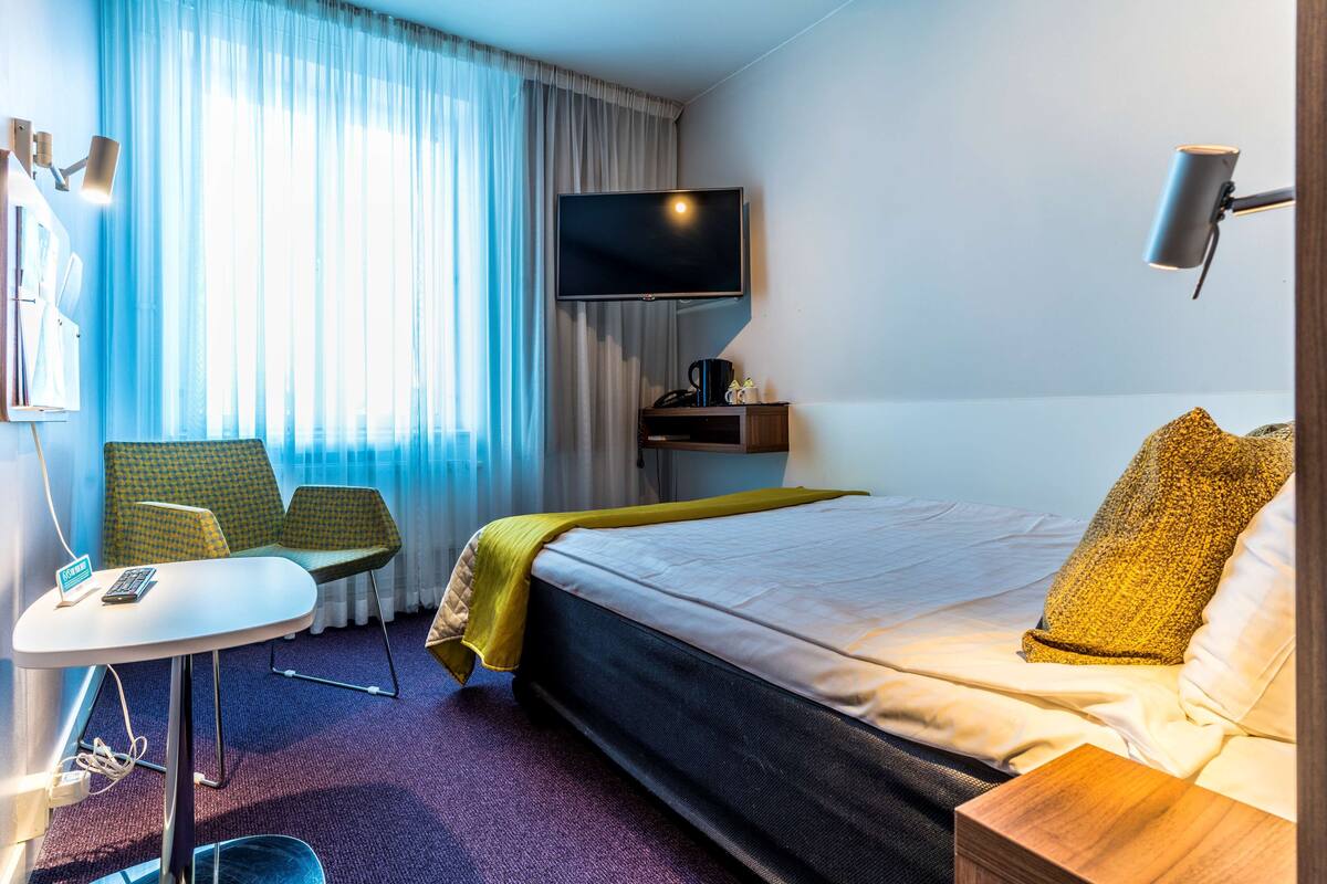 A hotel room with a bed, a desk with a chair, a TV, and a window with curtains.
