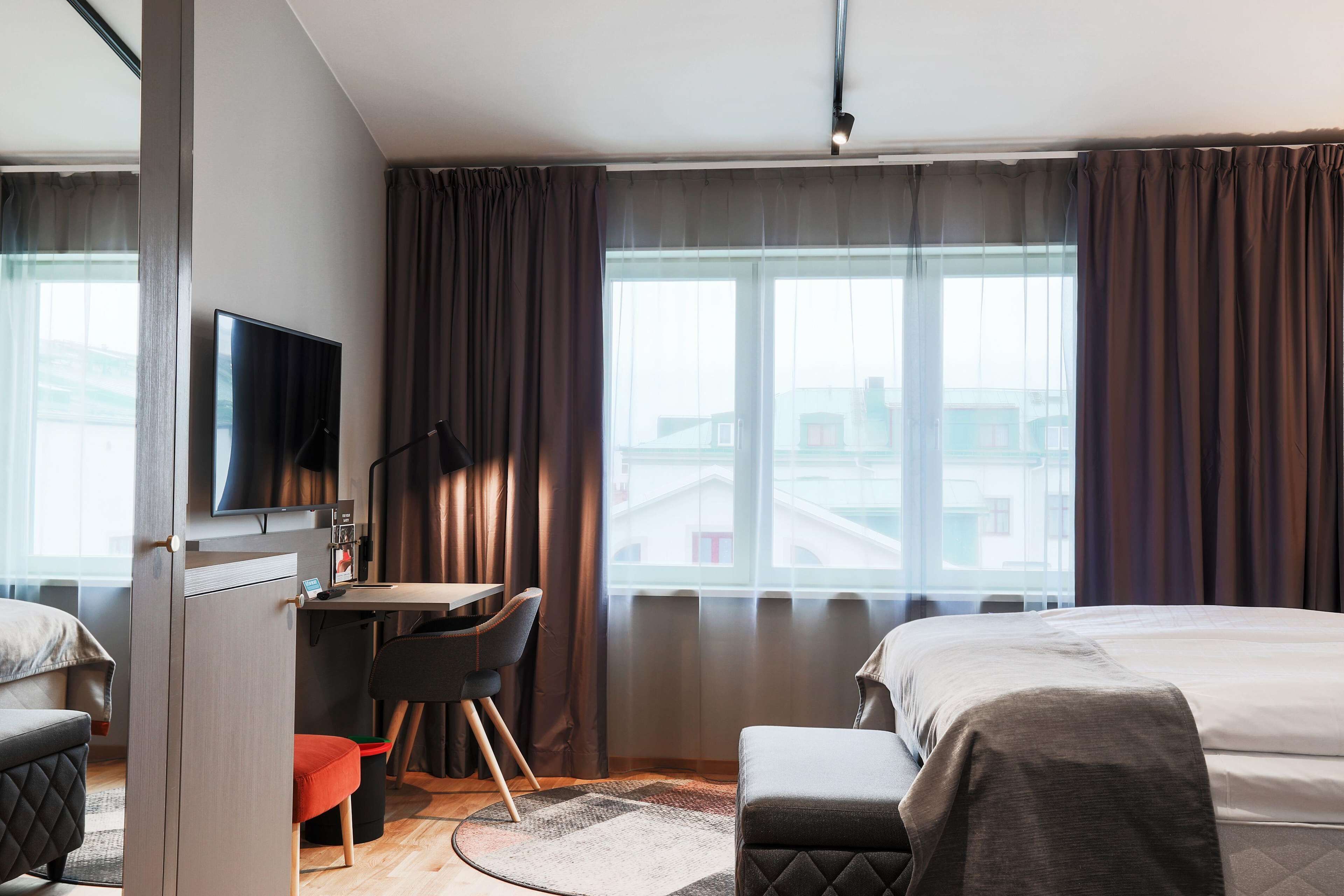 A hotel room with a bed, a desk with a chair, a television, and large windows with curtains.
