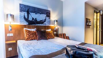 A hotel room with a large bed, a wooden headboard, two bedside lamps, a wall-mounted painting of a gondola, and a suitcase on the bed.