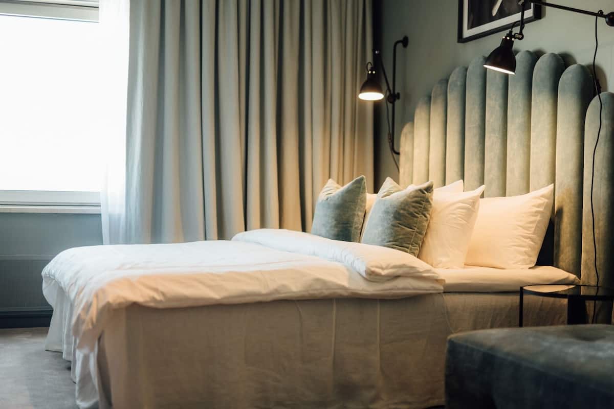 Cama bem arrumada com roupa de cama branca, luminária de cabeceira e radiador fixado na parede.