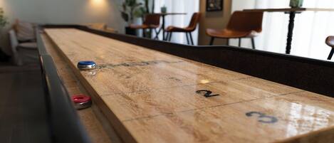 Ein Shuffleboard-Tisch mit nummerierten Feldern, einem blauen und roten Puck sowie einem unscharfen Hintergrund mit Stühlen und einem Tisch.