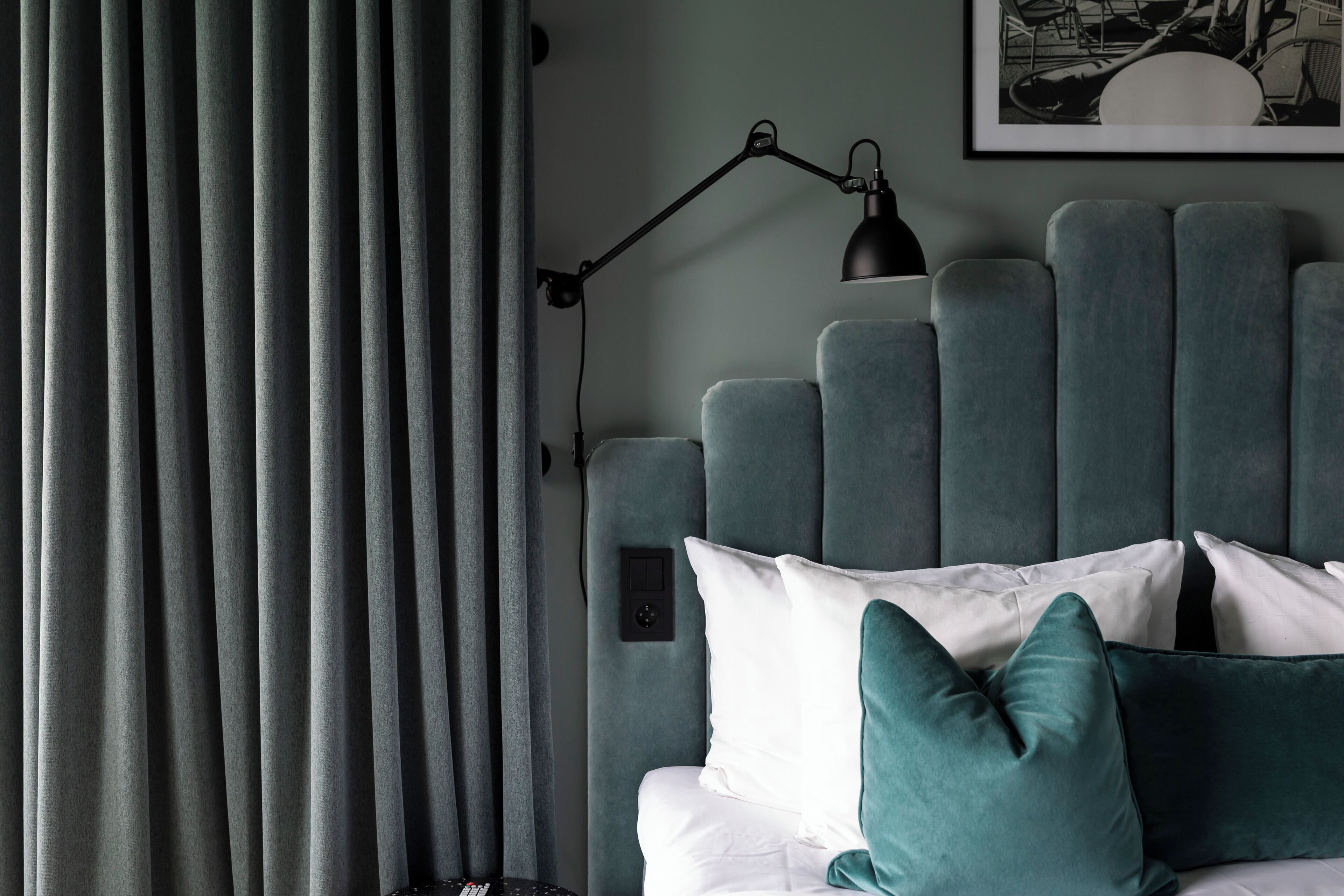 A bed with a headboard, white and teal pillows, a black desk lamp, and a framed picture on the wall.
