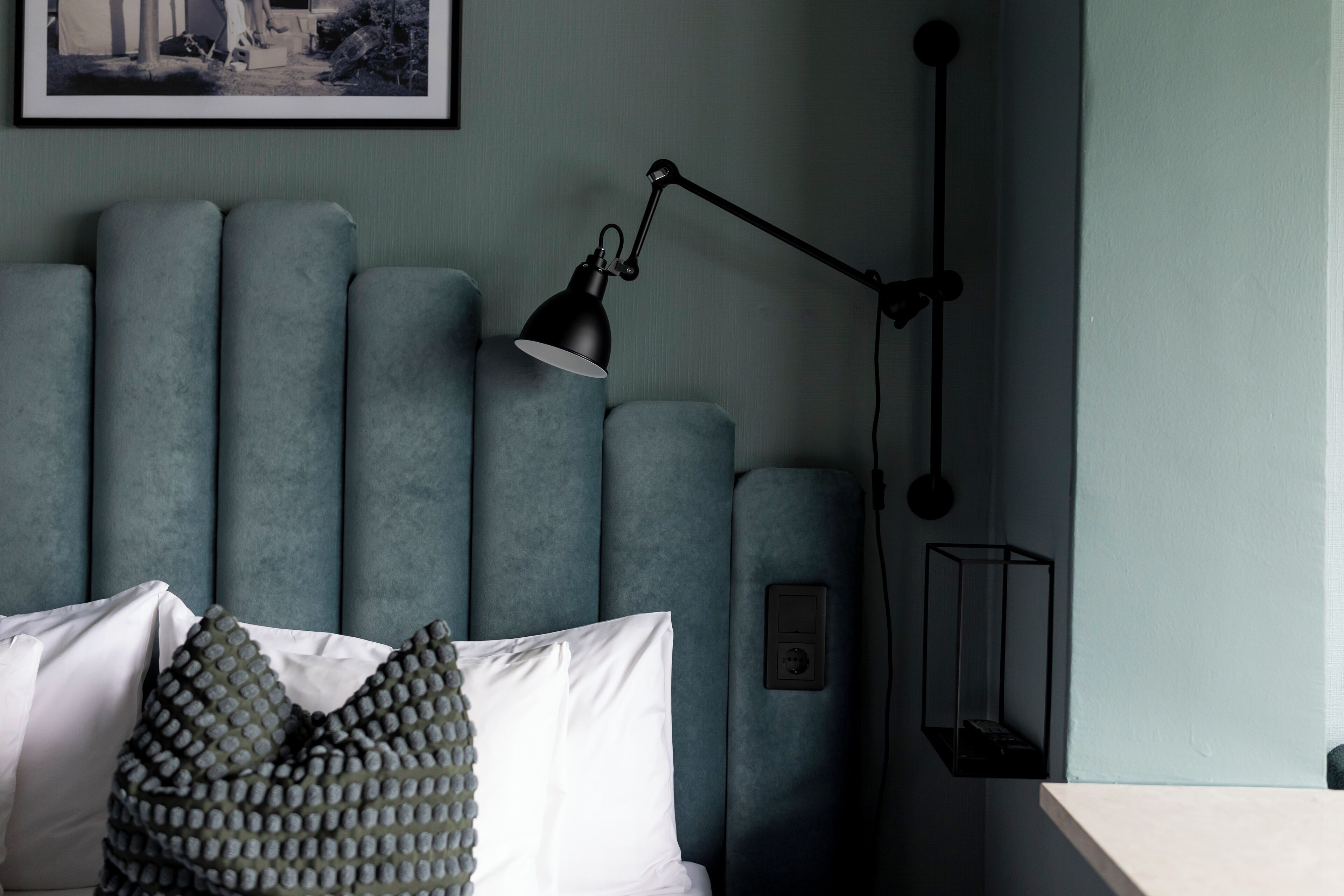 A modern bedroom with a grey upholstered headboard, a black desk lamp, and a framed picture on the wall.