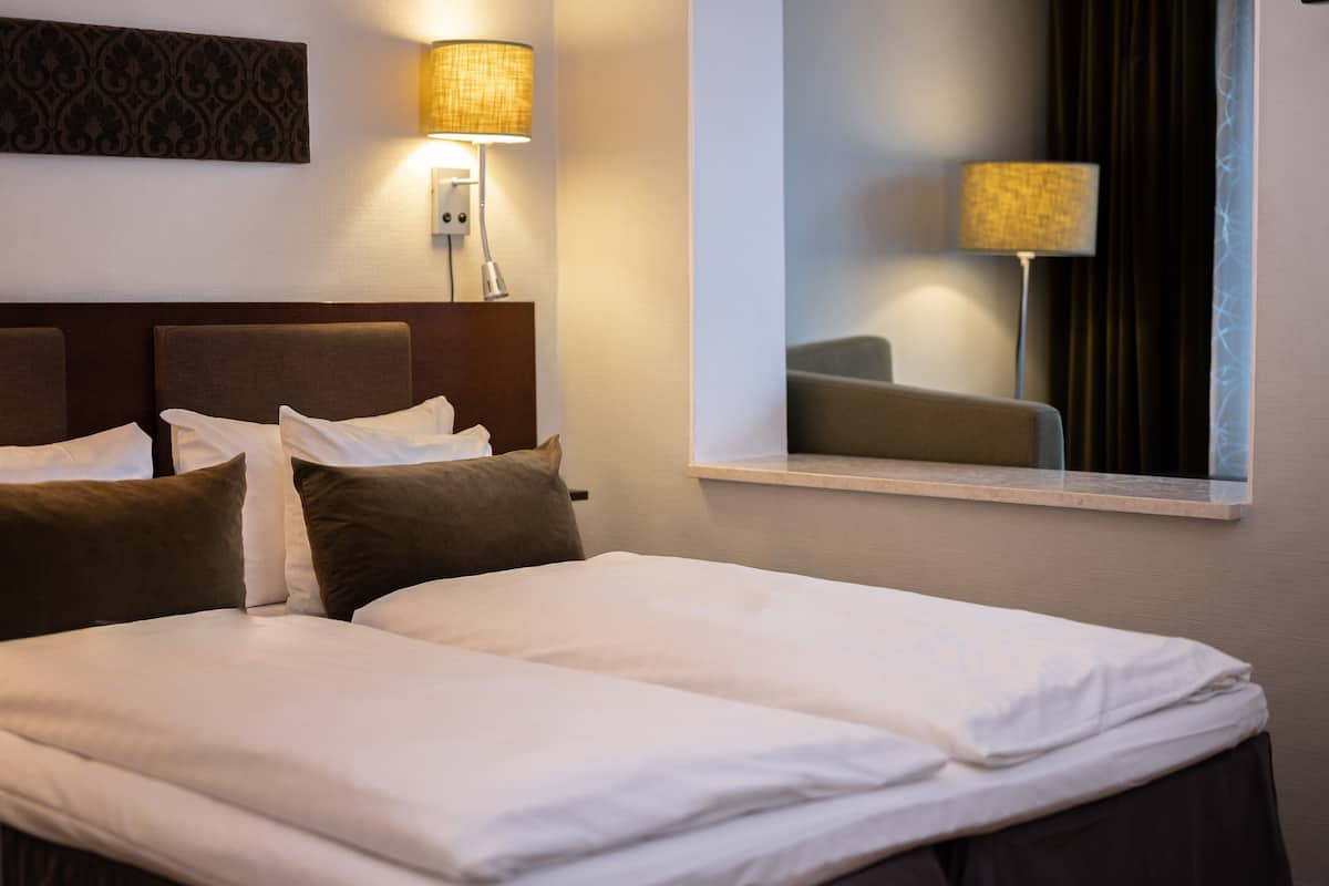 Quarto de hotel com cama, duas luminárias de cabeceira, um espelho e um painel decorativo fixado na parede.