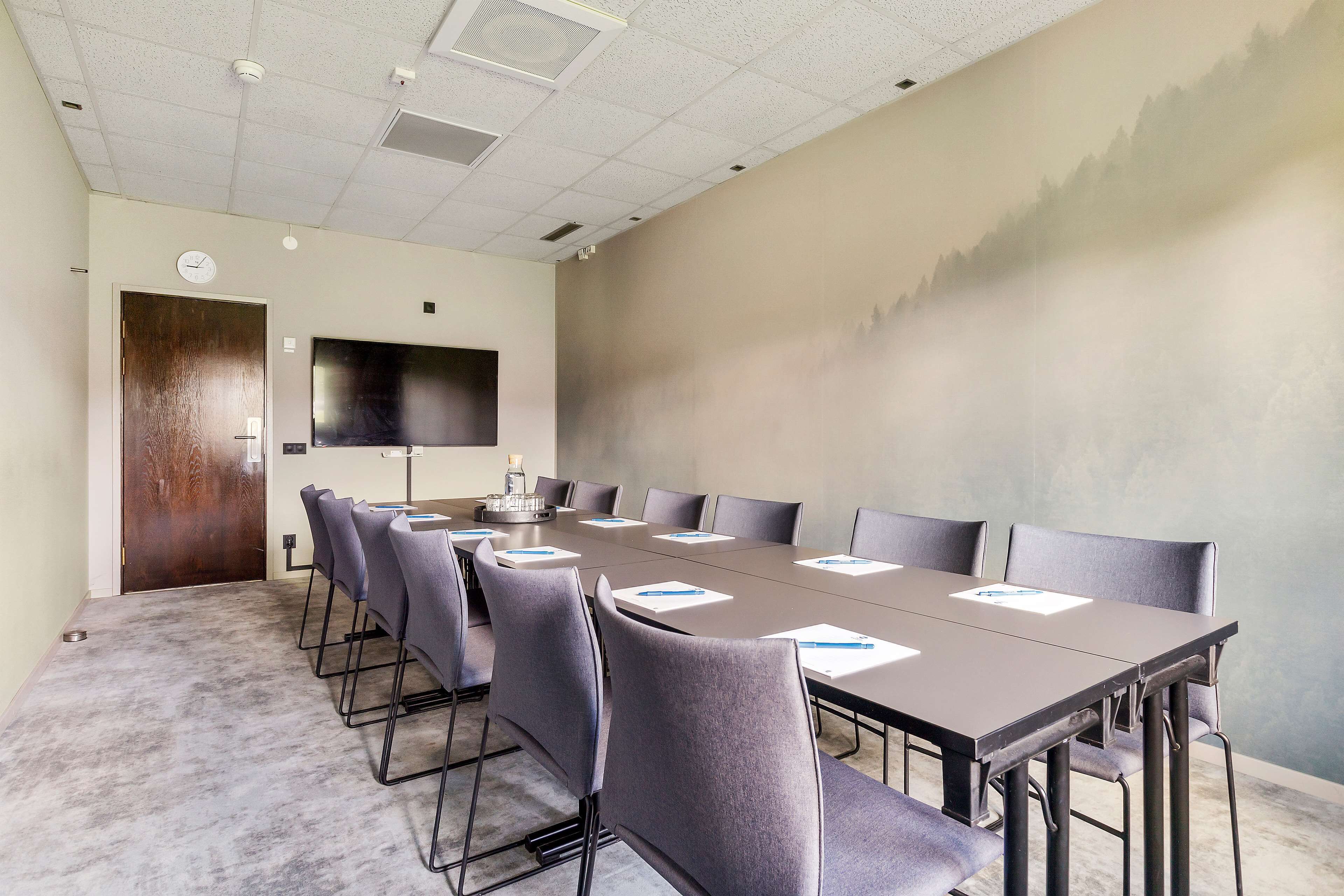 A conference room with a long table, grey chairs, a clock, and a TV on the wall.