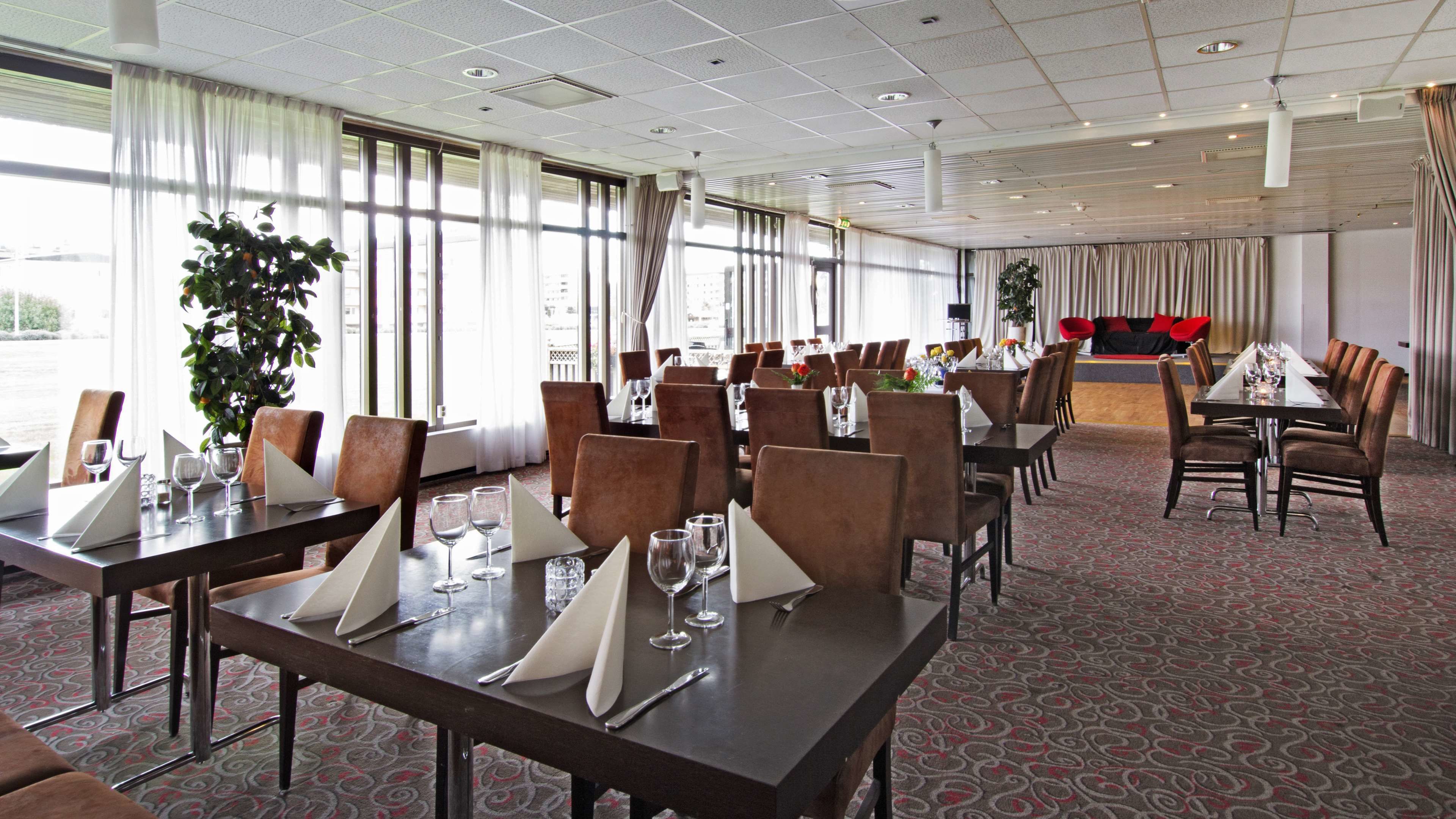 Une salle à manger d’hôtel avec de nombreuses tables dressées, de grandes fenêtres avec des rideaux et une scène où l’on aperçoit des chaises rouges à l’arrière-plan.