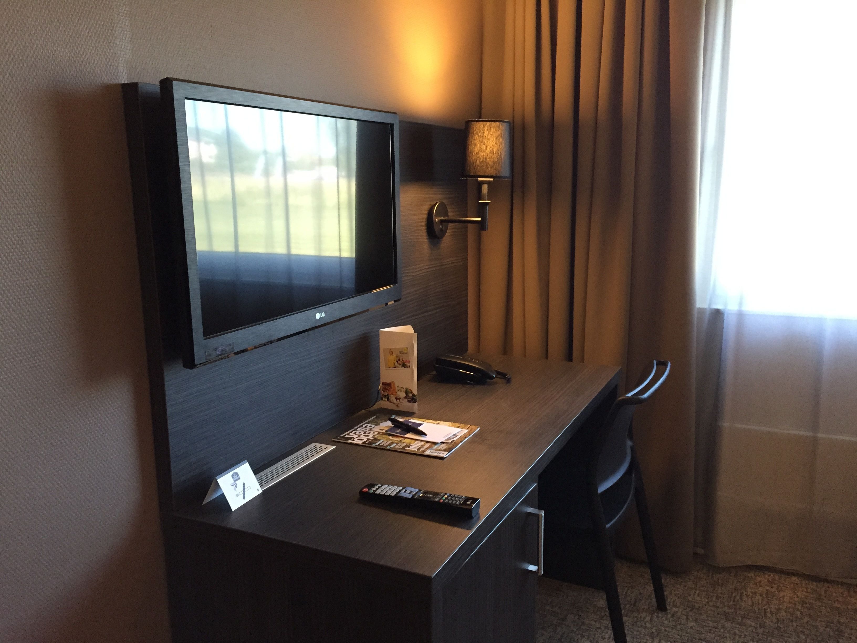 A hotel room with a flat-screen TV mounted on the wall, a desk with a remote control, a lamp, and a chair.