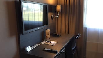 A hotel room with a flat-screen TV mounted on the wall, a desk with a remote control, a lamp, and a chair.