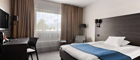 A hotel room with a bed, a desk, a chair, a TV, and a window with curtains.