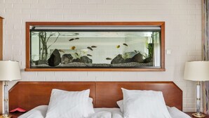 A hotel room with a bed, two bedside lamps, a large fish tank on the wall, and a white brick wall. - Best Western Hotel Scheele (Köping)