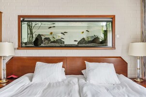 Habitación de hotel con cama, dos lámparas de noche, un acuario grande en la pared y una pared de ladrillos blancos.