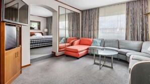 A modern hotel room with a sectional sofa, a glass coffee table, a flat-screen TV, a bed with checkered bedding, and large windows with curtains. - Best Western Hotel Scheele (Köping)