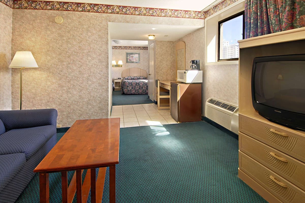 A hotel room with a sofa, a wooden table, a TV, a desk, a chair, a bed, and a window with curtains.
