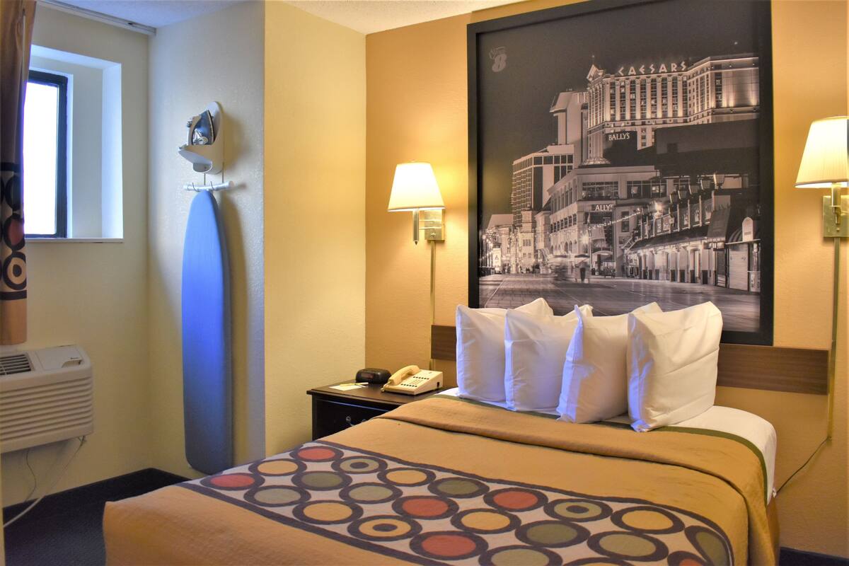 A hotel room with a bed, a nightstand, a telephone, a lamp, and a window.