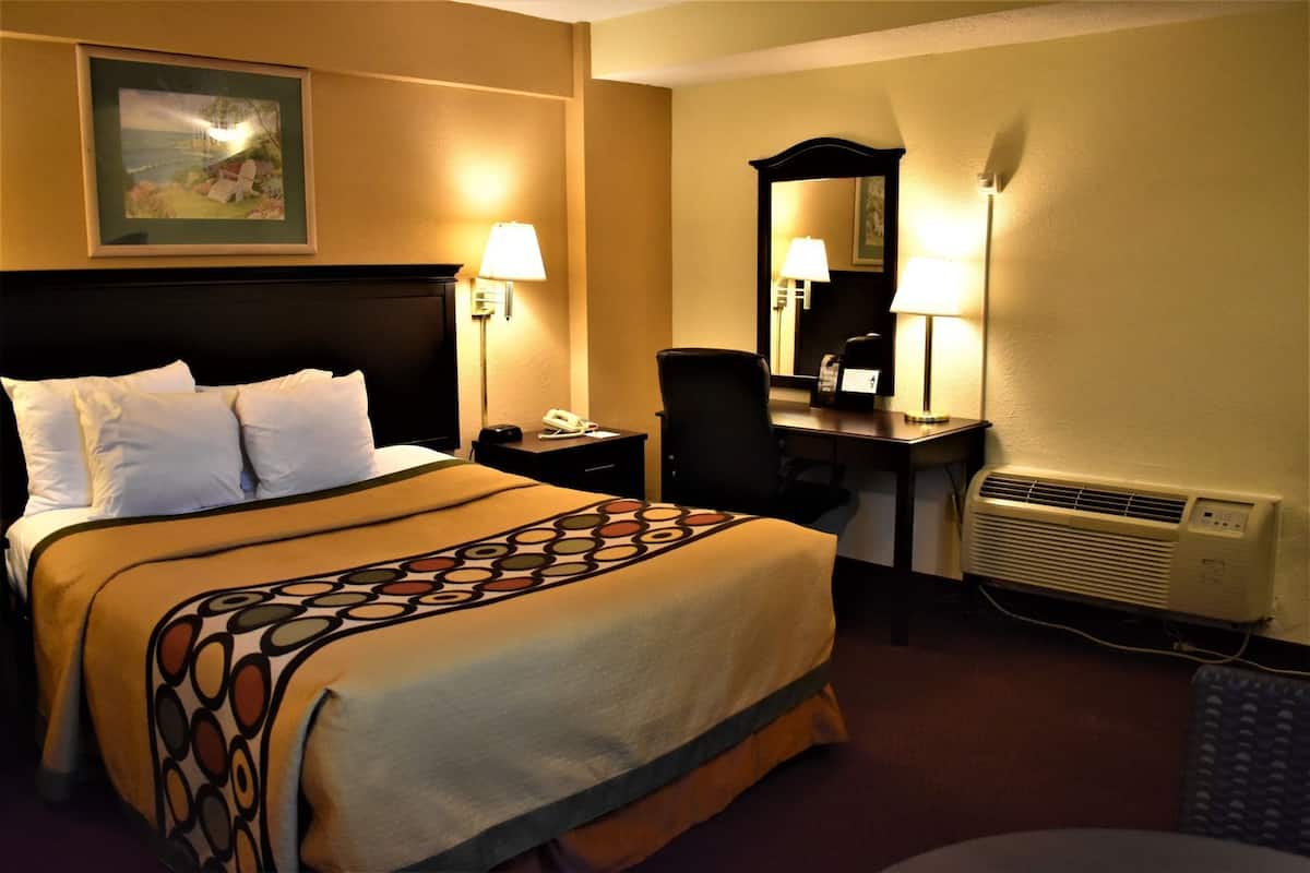 A hotel room with a bed, bedside lamps, a desk, a chair, a mirror, and a wall-mounted air conditioner.