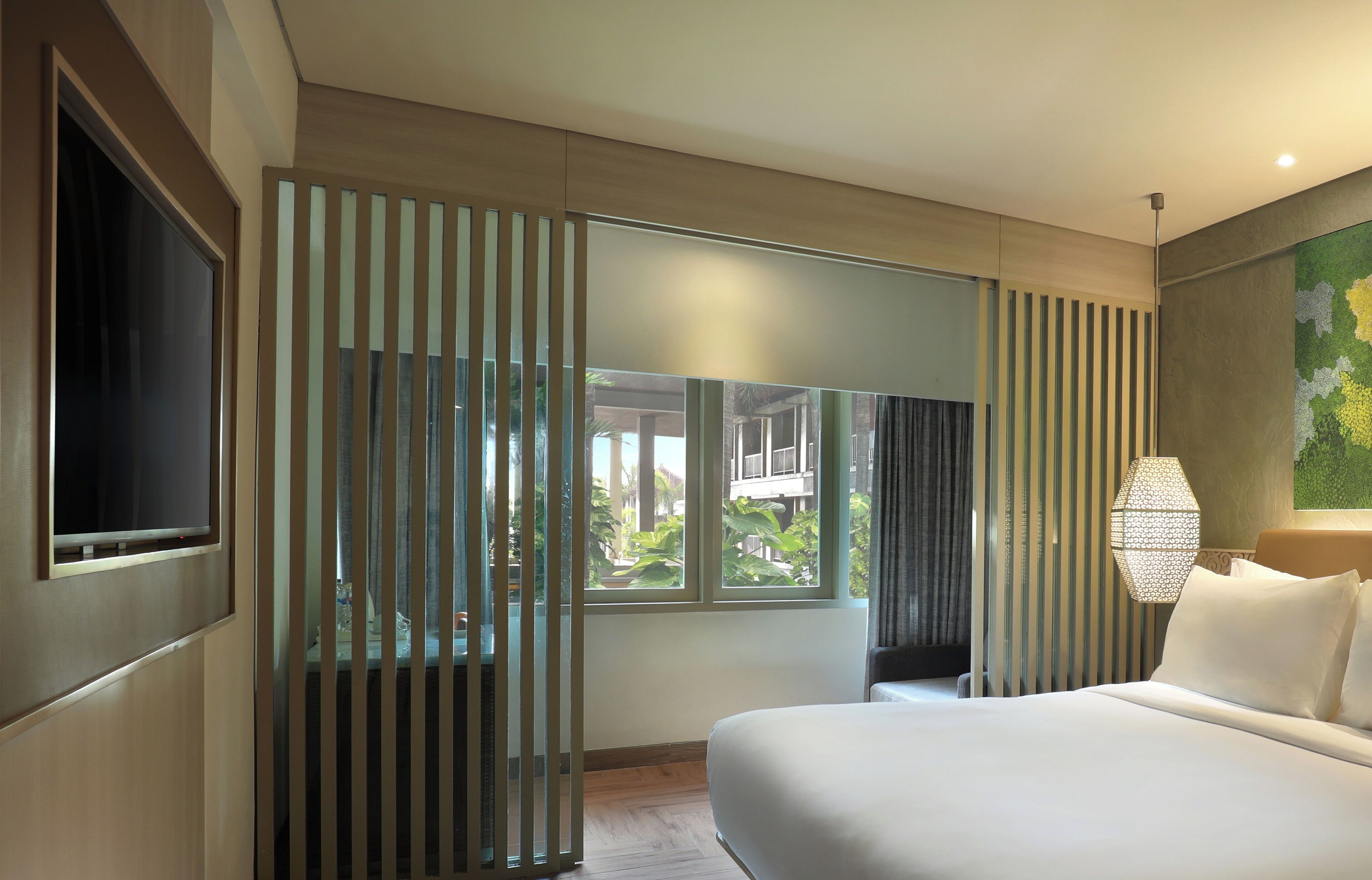 A modern hotel room with a large bed, a TV mounted on the wall, and a view of greenery outside.