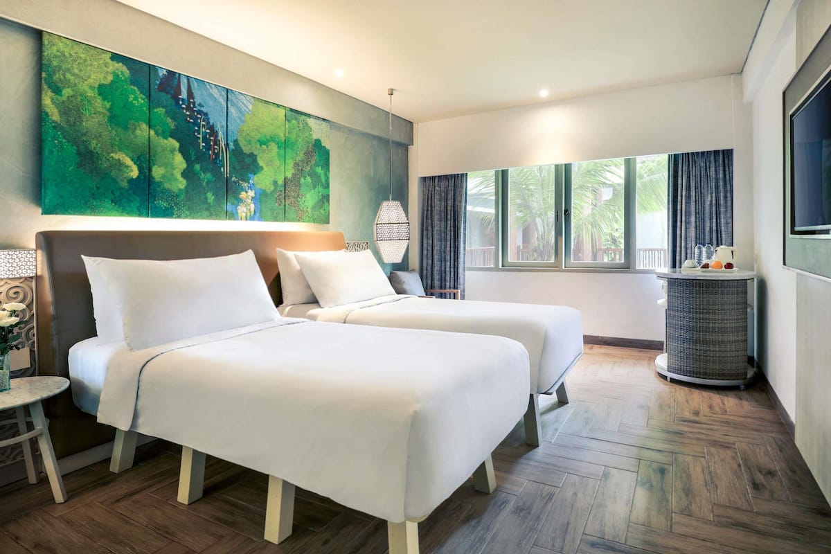 A hotel room with two beds, a large painting, a window with curtains, and a wicker side table.