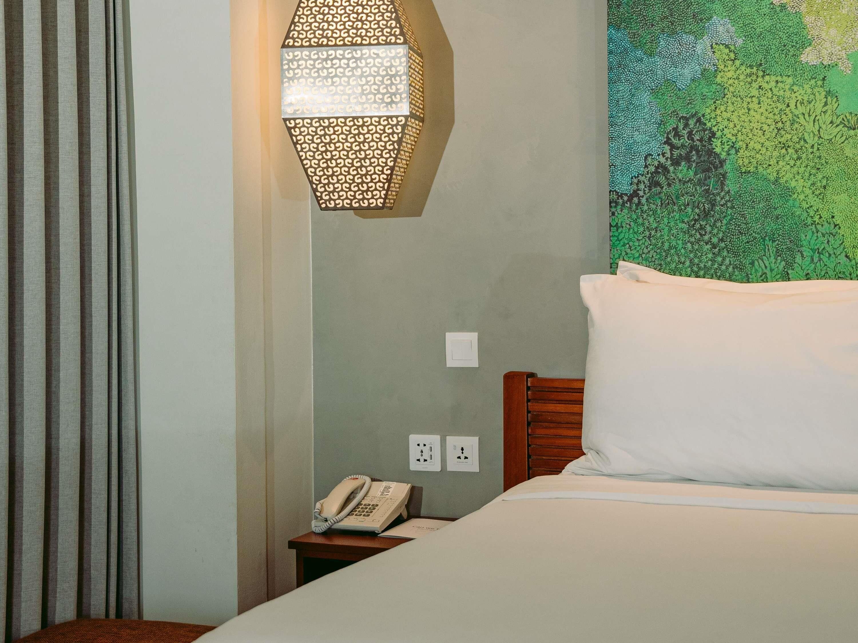 A hotel room with a bed, a bedside table with a telephone, a wall-mounted lamp, and a green patterned wall.